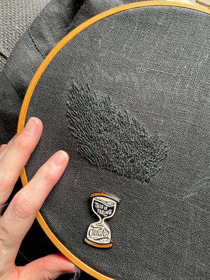 Lia’s fingers show a 25 by 3 inch wonky blob of black on black whipped backstitch that looks a bit like flames, but if you’ve been following this piece you’ll know it’s pubic hair. It’s definitely pubic hair. An hourglass-shaped needle minder that reads “this is taking forever” is attached to the cloth.