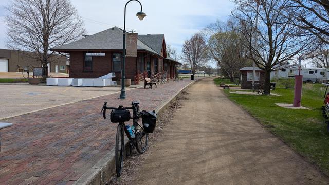 Dark brown Diverge bicycle leaning on the brick train platform in front of the Sparta Wisconsin vintage red brick train station with the crushed rock La Crosse River Trail on the right.