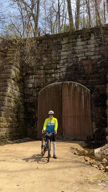 A man in a hi viz jacket and bicycle gear holding his Diverge bicycle on the Elroy - Sparta State Trail in front of the stone entrance to Tunnel 3 with the wooden doors closed and locked.