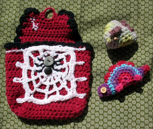 Red crochet pouch from the front with spiderweb granny square as decoration and 2 black buttons that were turned into a spider with some embroidery. Next to it a small triangle grey-ish crochet chicken with tiny wooden beads as eyes and a red-purple-blue crochet stegosaurus with wooden buttons as eyes, after tutorials by @sweetsofties . They rest on green flower-pattern fabric.