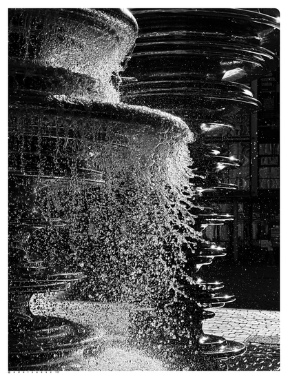 Schwarzweißfotografie: über silberfarbene glänzende Brunnenskulptur, unterschiedlich große kreisförmige Durchmesser, fällt Wasser spritzend nach unten, tausende kleine Wassertropfen