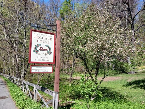 Photo of a red-bordered sign that reads "Louisa May Alcott's ORCHARD HOUSE Home of Little Women". In the middle of the sign is "Alcott" in a red script font between 2 curved branches of evergreens with 3 red berries each. Another smaller red-bordered sign below it reads "louisamayalcott.org" and below that "OPEN DAILY". 

The sign is hanging on a brown wooden post at the end of a gray, weathered wooden fence that separates the property from the sidewalk on the left. To the right of the sign is a flowering tree just beginning to bloom and grassy, wooded property.