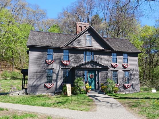 Photo of the front of Orchard House, Louisa May Alcott's home, where she wrote and set LITTLE WOMEN. It is a large, dark brown Colonial style house with 10 windows on the front. The green front door is adorned with a wreath. On each side of the front steps is a pot of many yellow daffodils in front of a pot of many yellow tulips. On each side of the front door, half-circle red-white-and-blue Americana bunting hangs below 2 top windows and 2 bottom windows.