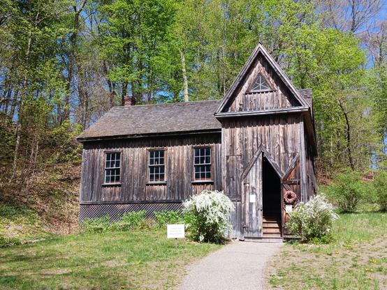 Photo of the Concord School of Philosophy, which was built just behind Orchard House. It is a weathered-looking brown wooden chapel style building with vertical siding. Louisa May Alcott's father used the building for meetings to share and discuss ideas.