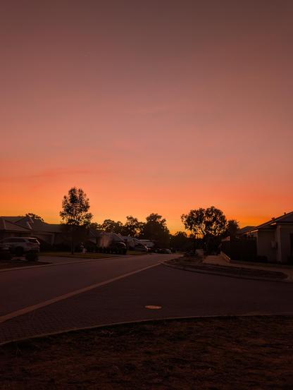 A suburban street at sunrise with houses on either side and trees silhouetted against an orange and pink sky.