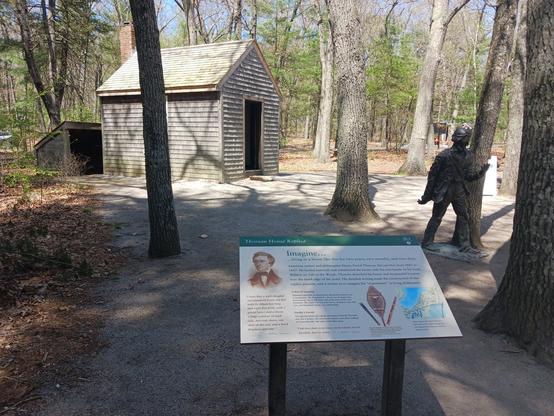 Photo of a replica of Henry David Thoreau's 10' x 15' one-room house. It is situated among trees and there's a small slanted-roof shed behind it. In the foreground is an informational sign about the house, and off to the right, between the sign and the house, is a statue of Thoreau.