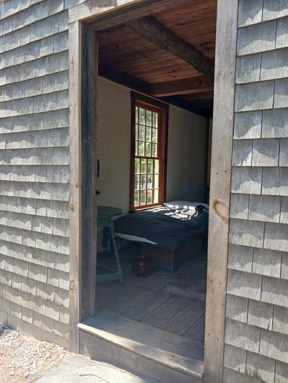 Photo looking in through the frame of the front door of the Thoreau house replica. There is a simple bed under a window in the middle of the left wall of the house.