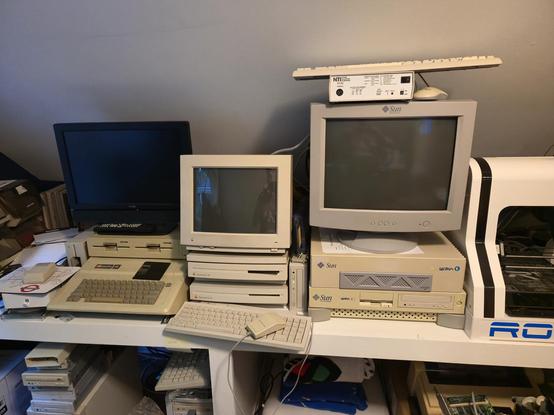 a picture of retro computers sitting on a builtin desk.  the right most stack has a Sun Ultra 1 sitting on a Sun Ultra 5 with a Sun CRT on top.  the KVM, keyboard, and mouse are on top of the monitor. the middle stack has two Mac LC III's sitting on top of each other with an apple 13 in monitor on top.  the keyboard for the Macs is sitting in front of the stacked Macs.  on the far left is an Apple IIe with a Duo disk and a flat panel monitor on top.