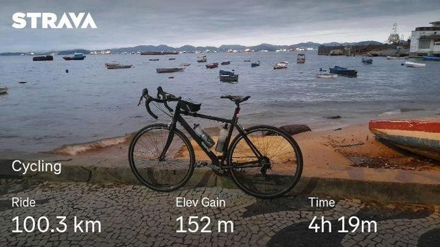 Road bike on a mosaic-tiled sidewalk by the bay shore in late evening. A small beach and moored boats on the water are visible in the background. Strava overlay showing 100.3 km biked in 4h 19m.