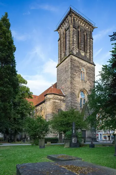 The image features a historical stone church with a prominent tower, surrounded by trees and greenery. The building has architectural details typical of medieval structures, such as arched windows and a textured facade. The scene is set against a bright blue sky, indicating a clear day. There are also gravestones visible in the foreground, suggesting that the area may include a cemetery.

Image Credits: Wikimedia / ChristianSchd / CC BY-SA 3.0