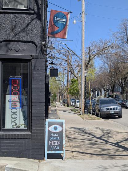 A daytime photo of a local bookstore from the side of the entrance. The lighted books sign can be seen through the window. Above the door the shop sign and below it is a chalkboard with an eye and the phrase Make Orwell Fiction Again.