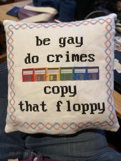 a little pillow with cross-stitch on the front. it says “be gay do crimes”, then there are six 3.5” floppy disks in the gay rainbow colors, and below that it says “copy that floppy”. the border is pink and blue, and the background is white; it’s meant to evoke the trans flag, but I’m not sure that really landed.