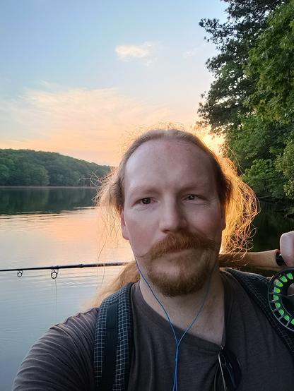 A handsome angler with a fly fishing rod over his shoulder. Behind him is an expanse of lake with a green hill on the opposite shore. The sun is setting behind his head, and the golden light catches in his hair to form a sort of halo. He has big moustache energy.