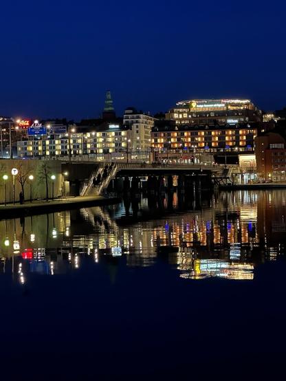 a system of bridges, and behind them, a cluster of lighted buildings, reflecting in a body of water at around dusk.