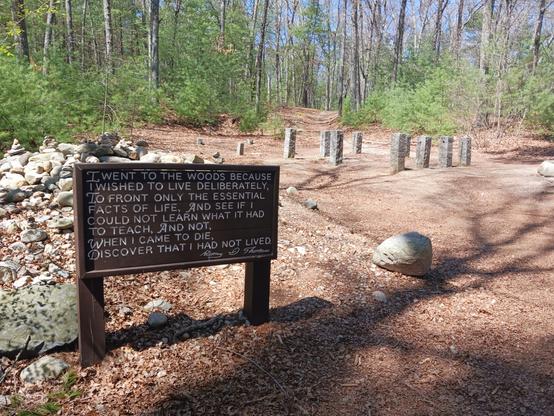 Photo of the original site of Henry David Thoreau's house. In the foreground is a large brown wooden sign with the following quote in carved lettering painted white: 

"I went to the woods because I wished to live deliberately, to front only the essential facts of life, and see if I could not learn what it had to teach, and not, when I came to die, discover that I had not lived."

Below the quote is the carved signature, painted white, of "Henry D Thoreau".

Behind the sign is a rock cairn made of stones placed by visitors as a natural memorial to Thoreau. 

In the background are granite pillars that mark the original site of Thoreau's 10' x 15' house. Beyond the site are woods.