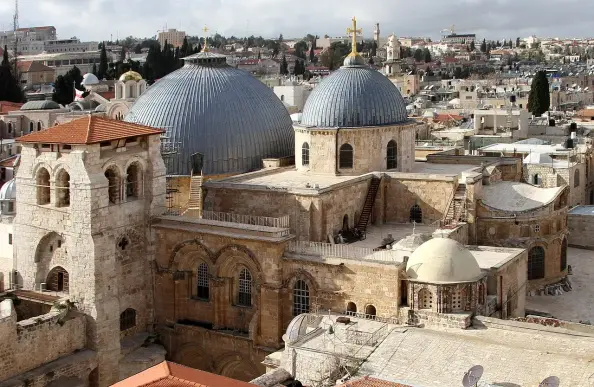 The image shows a historical complex with domed structures and stone architecture, likely featuring religious significance. It appears to be a view of a church or monastery, characterized by its distinct domes and surrounding buildings, perhaps set in an ancient city like Jerusalem. The landscape includes stone walls and rooftops, typical of the area's architectural style.

Image Credits: Wikimedia / Gerd Eichmann / CC BY-SA 4.0