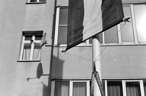 The black and white image shows a close-up of a building façade with windows, a satellite dish casting a clear shadow on the wall, and a flagpole bearing the national flag of Austria secured with ropes.