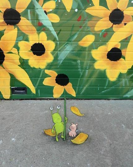 On a brick wall is painted orange flowers on a mural on a green background.

 David Zinn has painted an alien and a small pig holding a stalk, with loose petals, on the concrete floor in front