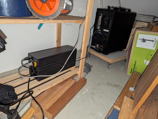 Ein schwarzer länglicher Kasten auf dem untersten Fach eines Holzkellerregals ist mit einem schwarzen PC verbunden. Der steht auf einem Rollbrett daneben.