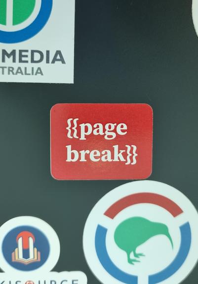 A sticker on a laptop lid, red with '{{page break}}' in white text, with parts of other stickers visible around it.