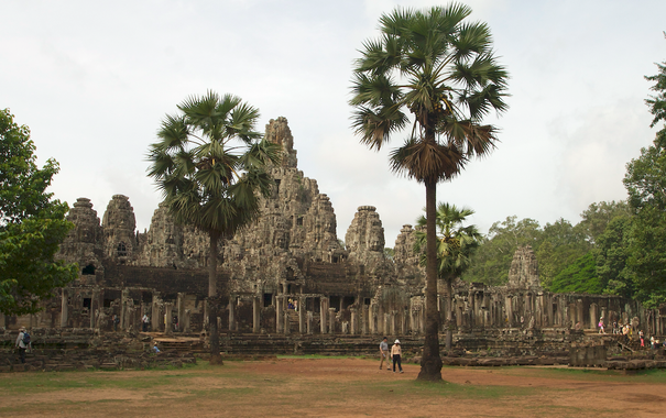 part of Angkor Wat - a large structure, resembling an intricately carved wall with faces and varied stones, can be seen across a field, behind several palm trees and under a grey, hazy sky