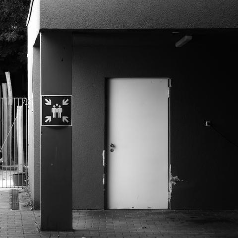 Ein quadratisches Schild an einer Betonsäule. Darauf sind piktografisch Menschen abgebiildet und aus jeder Ecke des Bildes ragt ein Pfeil auf diese Menschen. Das Foto ist in Schwarzweiß.
