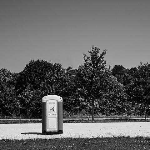 Eine sogenannte Baustellentoilette. An der Tür steht T0i T0i. Dieses Wc steht einsam auf einem breiten Weg dutch eine Parklandschaft.