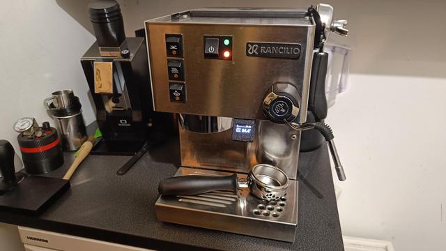 Meine Siebträgermaschine, man sieht ein kleines Display, das die Temperatur anzeigt. Im Hintergrund diverse okkulte Geräte zur Herstellung von Kaffee: ein Tamper, ein 3d-gedrucktes WDT Tool (zum Verteilen des Mehls im Sieb), eine Mühle mit Single-Dosing-Aufsatz usw.