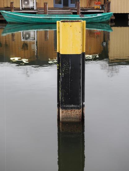 The image shows a black and yellow bollard in the water, likely used for mooring boats. In the background, there's a dock with a small green boat and some wooden structures. The water reflects the bollard and parts of the dock area.