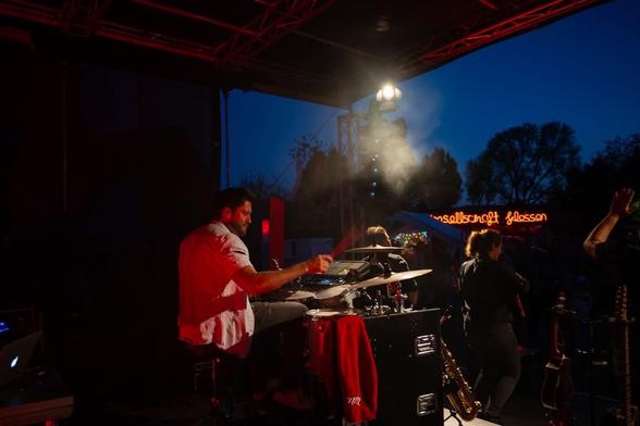 Ein Schlagzeuger während eines Konzerts. Im Hintergrund sind andere Musiker zu sehen, sowie Bäume.