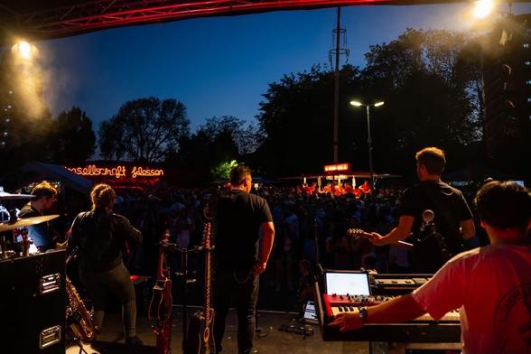 Aus dem hinteren Bereich einer Bühne fotografiert. Zu sehen sind die Musiker von hinten sowie ein voller Dorfplatz. Einen Getränkestamd, sowie Bäume sind im Hintergrund zu sehen.
