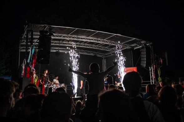 Blick aus dem Publikum auf eine Bühne, vorne sehen wir einen Jungen der auf den Schultern seines Vaters sitzt und die Arme ausgebreitet hat. Dahinter die Bühne mit den Musikern , sowie 2 kleine Feuerwerke die auf der Bühne gezündet werden.