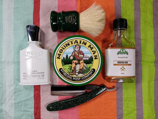 (Le Chat Mistral description)

This image shows a collection of men's grooming items arranged on a colourful striped fabric. There's a white bottle of Creed Silver Mountain Water fragrance on the left, with a green shaving brush holder above it. In the centre is a round tin of Stirling Soap Company's Mountain Man shaving soap, featuring an illustration of a bearded man in outdoor gear. To the right is a small bottle of Stirling's Mountain Man aftershave splash. Below these items is a straight razor with a dark handle, lying open. The overall scene is vibrant and well-organised, highlighting classic shaving essentials.