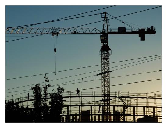 Foto im Querformat. Die Silhouette einer Baustelle kurz vor Sonnenuntergang. Der obere Teil eines runden Baugerüsts, davor teilweise Baumspitzen. Zwei Menschen laufen oben auf dem Gerüst. Dahinter ist ein Kran, der seitlich zur Kamera steht. Der Himmel ist blau, weiter unten gelblich. Viele Leitungen verlaufen durchs Bild.
