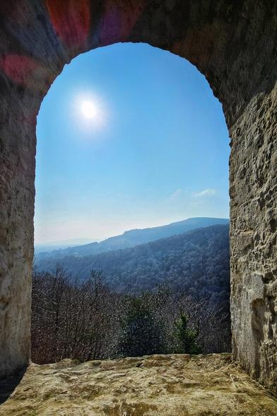 A view of a forested valley and distant hills seen through an arched stone window opening. The sun shines brightly in the clear blue sky.