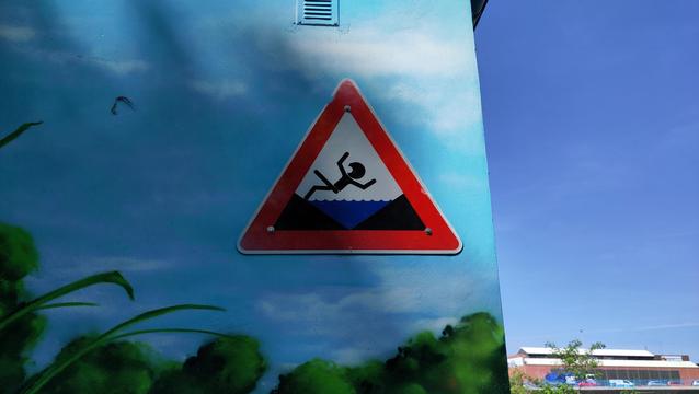 Dreieckiges Warnschild an einer Gebäudewand. Ein stilisiertes, sehr abstraktes Männchen fällt rückwärts ins Wasser. Der Mund ist aufgerissen, die Arme rudern in der Luft.
