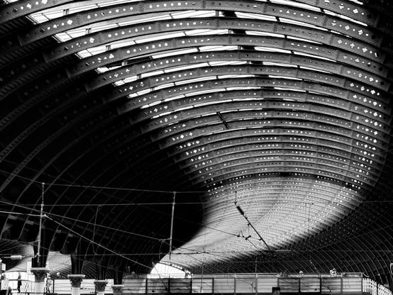 This image captures the interior of a large, arched structure, likely a train station or industrial building. The ceiling is composed of repeating metal beams and panels, creating a rhythmic pattern that curves overhead. The space is vast and open, with a sense of grandeur. In the foreground, there are some structural elements and possibly equipment, suggesting a functional or industrial setting. The image is in black and white, emphasizing the contrast and geometric shapes. The overall atmosphere is one of architectural elegance and industrial strength.