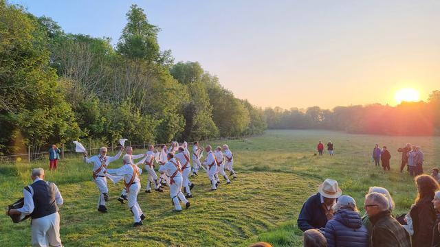 A bunch of men in white, with colourful sashes and white handkerchiefs, dance in a scythed circle of a meadow, while the sun rises from behind the trees.