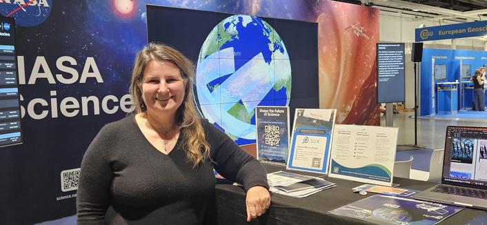 Woman stands in front of table displaying Science Explorer (SciX) literature with NASA hyperwall in back displaying visualization of earth science data