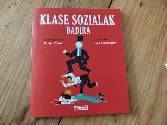 Egurrezko mahaian liburu gorri bat, klase zapalkuntzaren ilustrazio batekin, pertsonai boterearen arabera besteak zapaltzen literalki, eta Klase sozialak ere badira tituluarekin letra txurietan.