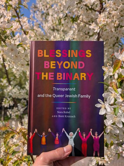 Holding the essay collection "Blessings beyond the Binary" in front of a tree with white blossoms