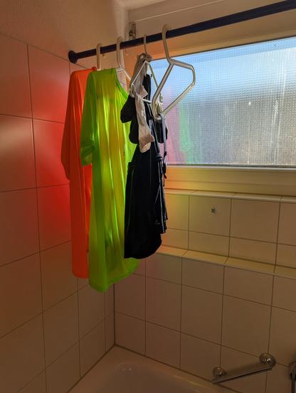Laufkleidung hängt auf Bügeln an einer mittig über der Badewanne angebrachten Duschvorhangstange ohne Duschvorhang.