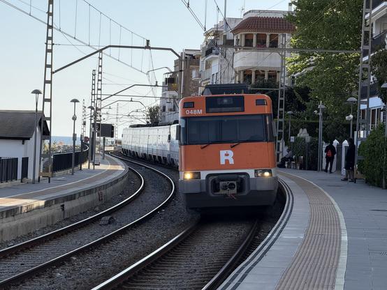 Tren entrando en curva en el lado derecho de la foto, es una estación en curva con dos andenes haciendo una ese. Tiene los focos encendidos en cortas.

A la derecha se ven unos edificios con algún árbol o matojo y a la derecha parte de una caseta y en el fondo el mar.

Se ve el cielo que aún se ve claro pero está empezando a atardecer.