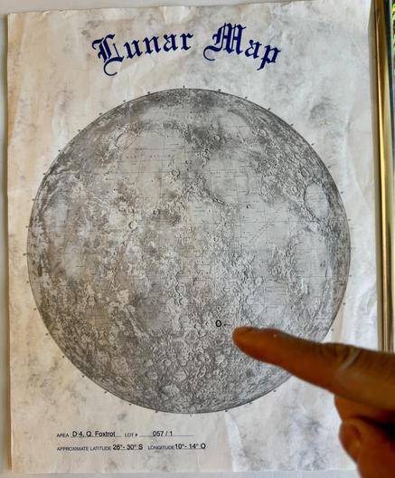 A lunar map featuring detailed topographical features of the moon. The image has a finger pointing at a specific location marked with a small 'O'. The map includes labels and coordinates in the bottom section.