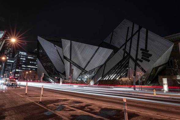 Long-exposure night photo of the Royal Ontario Museum in Toronto, showing the modern crystal-inspired architecture lit against the dark sky, with light trails from passing traffic in the foreground.