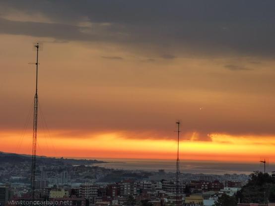 Amanecer. Skyline en primer término y mar, sobre el una franja de nubes anaranjadas que se van suavizando hasta el gris en la zona superior. Dos largas antenas cruzan la foto a lo lejos