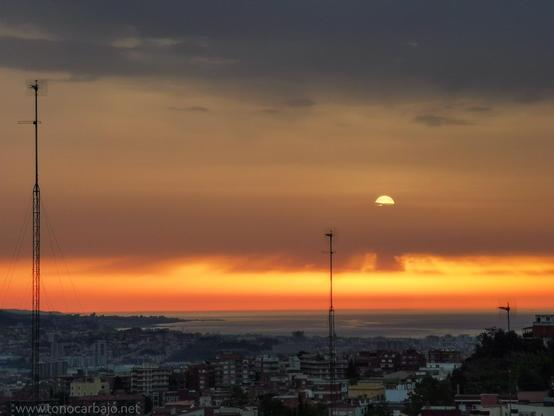 Amanecer. Skyline en primer término y mar, sobre el una franja de nubes anaranjadas que se van suavizando hasta el gris en la zona superior. Un tercio de sol aparece naranja entre las nubes. Dos largas antenas cruzan la foto a lo lejos