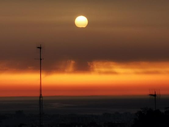 Amanecer. Skyline en primer término y mar, sobre el una franja de nubes anaranjadas que se van suavizando hasta el gris en la zona superior. El sol aparece naranja entre las nubes. Una larga antena cruzan la foto a lo lejos. El plano es un poco más cerrado que las dos anteriores fotos.