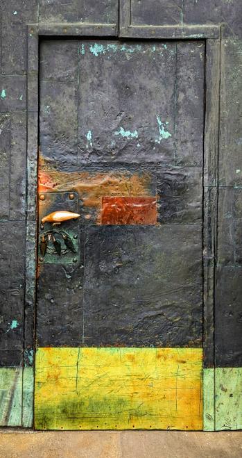 A weathered, dark metal door with a bright yellow lower panel. The metal shows signs of age and oxidation, with patches of turquoise and orange. A decorative metal handle and escutcheon are visible on the left side. The door is set within a frame of similar dark, textured metal.