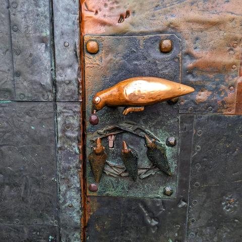 The image shows a close-up of a weathered, dark metal door. The metal surface exhibits signs of age and oxidation, with patches of turquoise and orange. A decorative metal handle and escutcheon, featuring a bird-shaped handle and a design of birds on branches, are visible on the left side of the door. When using the door handle, it looks like the bird that forms the handle is feeding the smaller birds below.